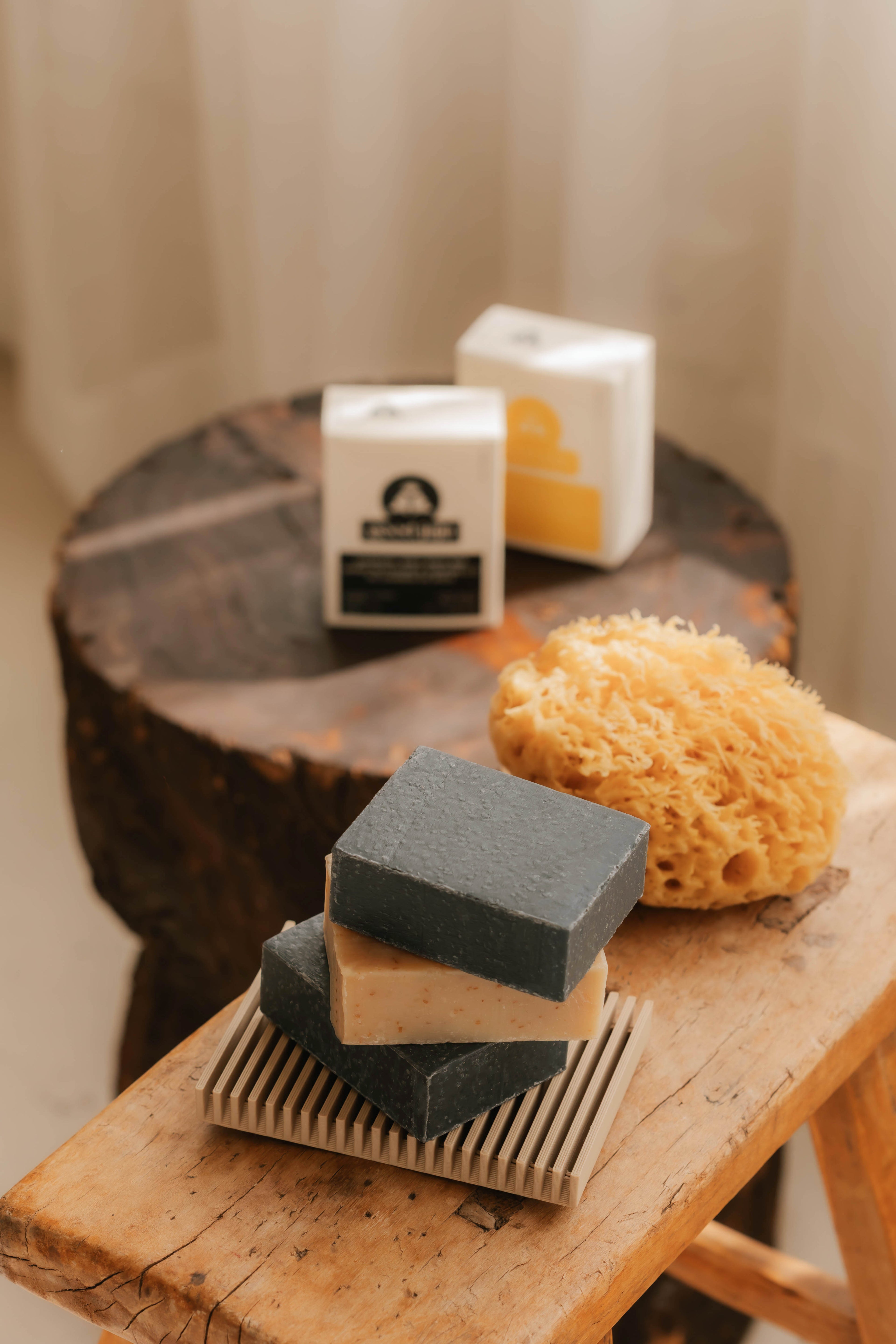 Three bars of soap on a wooden stand with a loofah and two containers in the background.