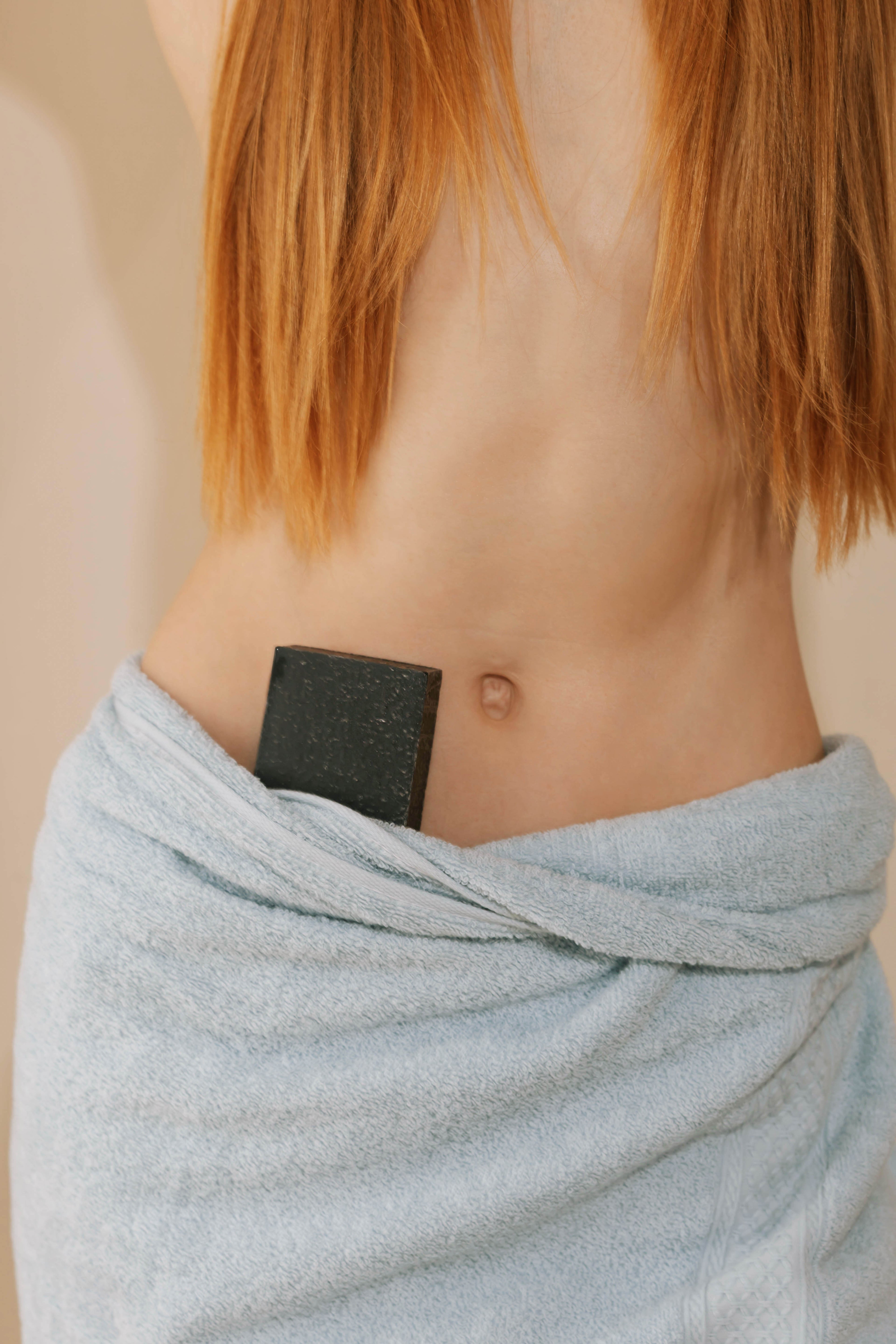 Black bar of charcoal soap tucked into the waistband of a blue towel on a model with light red-brown hair.