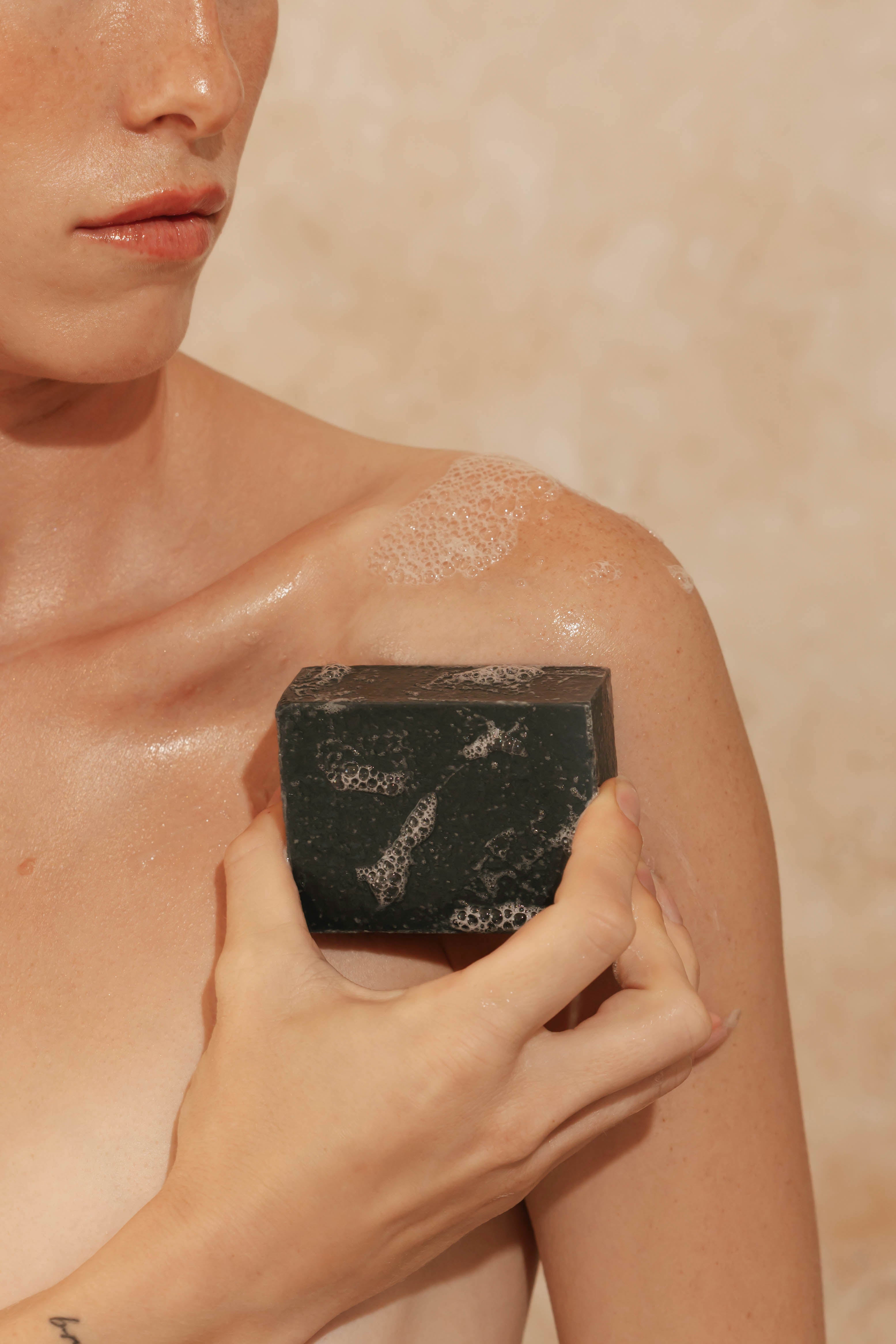 Person applying a black charcoal body wash bar to their shoulder against a beige background